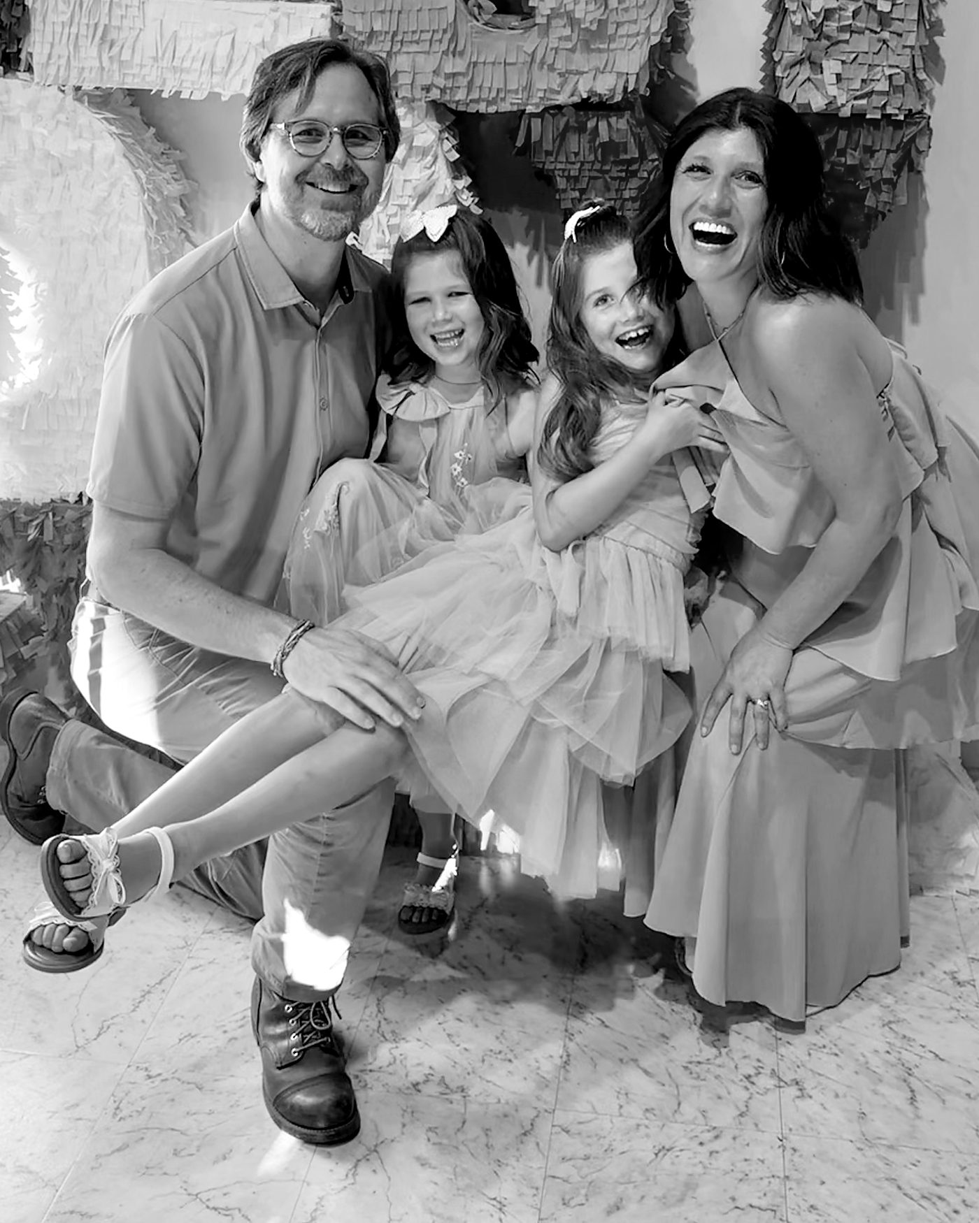 Black and white photo of David Dewese kneeling with his wife and two daughters, everyone laughing, in front of giant paper letters.
